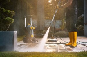 Pressure Washing. A worker in yellow boots uses a pressure washer to clean a patio area under bright sunlight, showcasing effective outdoor maintenance.