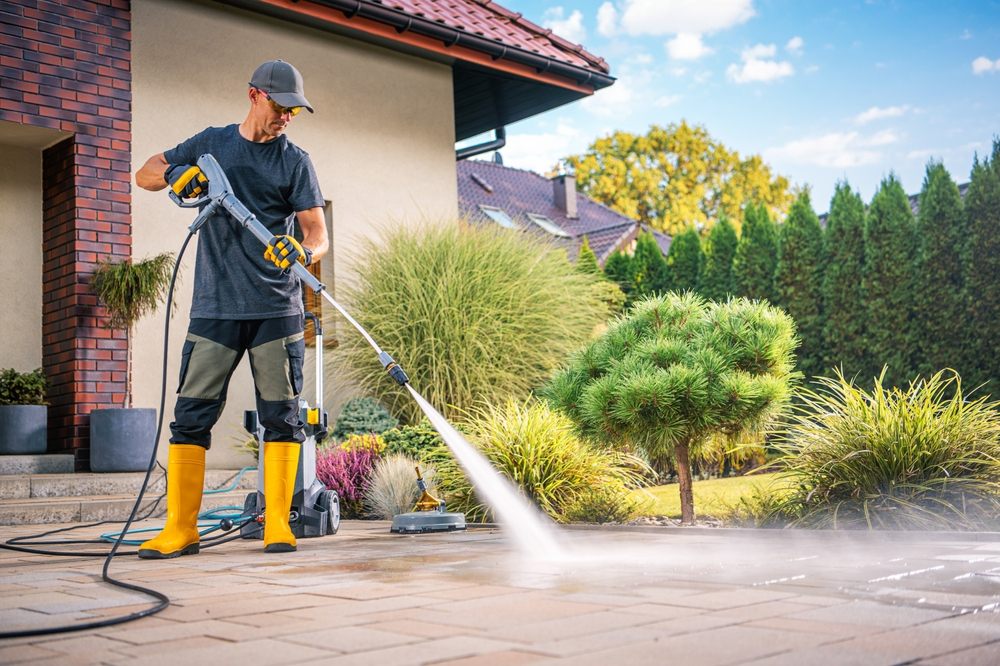 Why Vancouver Residents Trust Us for Pressure Washing Services. A man is using a power washer to clean the patio in a beautiful garden filled with greenery and sunshine.