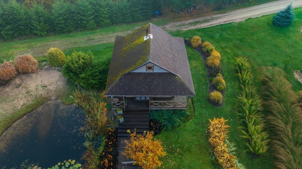 Roof Moss Removal & Treatment: Protecting Your Home the Right Way. Aerial image of a wooden house with a moss covered roof, surrounded by autumn trees, a pond, and a dirt road bordered by shrubs.
